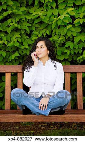 Young Colombian woman with long black hair white shirt and blue jeans, sits cross-legged on a wooden bench while talking on a mobile phone. She is looking off to her right. View Large Photo Image Stock Photo - Young Colombian woman with long black hair white shirt and blue jeans, sits cross-legged on a wooden bench while talking on a mobile phone. She is looking off to her right.. Fotosearch
