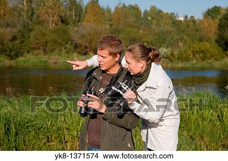 Picture - Young couple birdwatching with binoculars by river water pointing away. Fotosearch