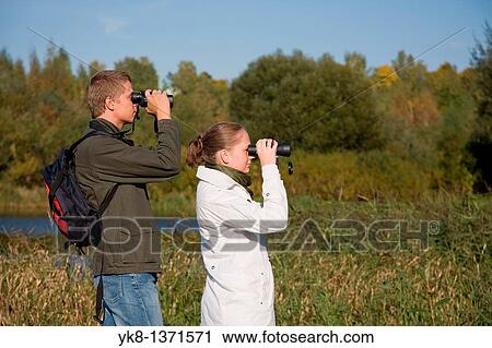 Stock Image - Young couple birdwatching with binoculars by river water. Fotosearch