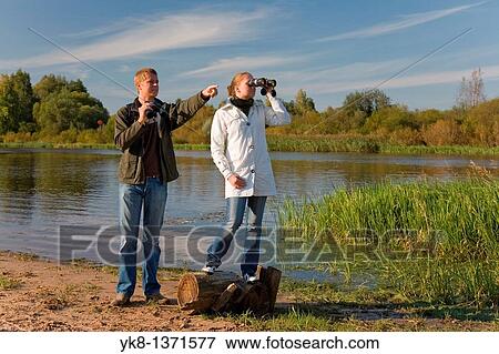 Stock Photo - Young couple birdwatching with binoculars by river water pointing away. Fotosearch