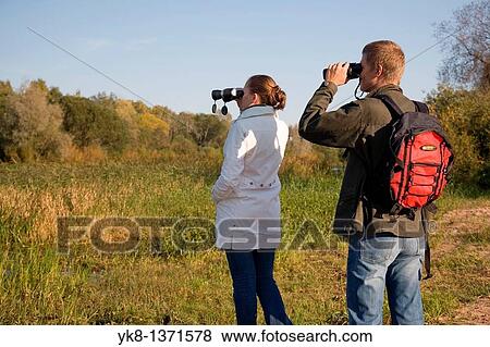 Stock Photo - Young couple birdwatching with binoculars by river water. Fotosearch