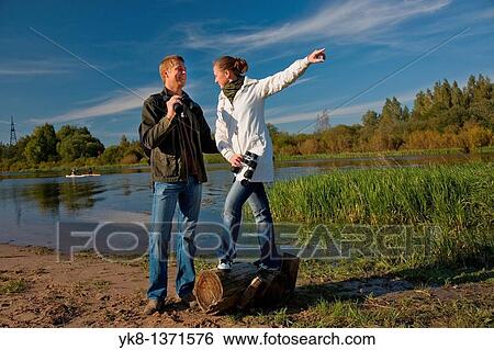 Stock Photograph - Young couple birdwatching with binoculars by river water pointing away. Fotosearch