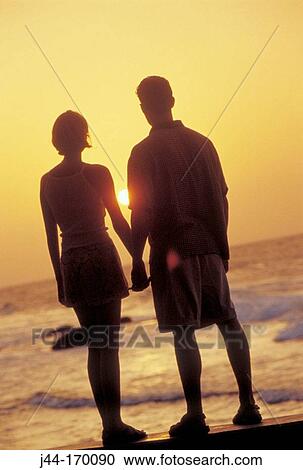 Stock Image - Young couple holding hands while looking the sunset at the beach, Venezuela. Fotosearch