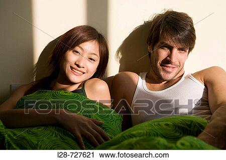 Stock Image - Young couple lying on bed, smiling, portrait. Fotosearch