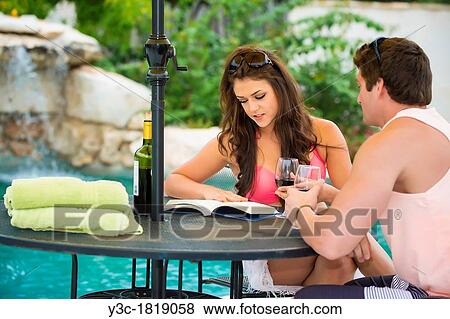 Young couple sitting at a table in a vacation resort's swimming pool area reading and enjoying wine View Large Photo Image Stock Photo - Young couple sitting at a table in a vacation resort's swimming pool area reading and enjoying wine. Fotosearch