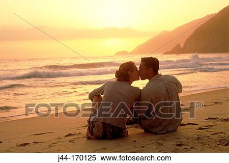 Stock Photography - Young couple sitting on sand at the beach kissing at sun set. Fotosearch