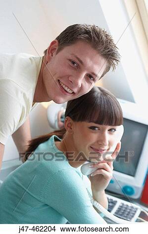 young couple sitting together in the office shes on the phone both smiling at the camera View Large Photo Image Picture - young couple sitting together in the office shes on the phone both smiling at the camera. Fotosearch