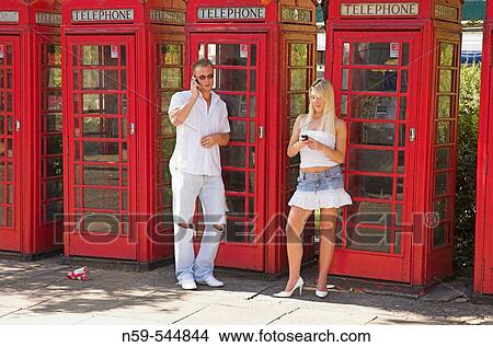Picture - Young couple using mobile phones on street next to row of British public phone boxes.. Fotosearch