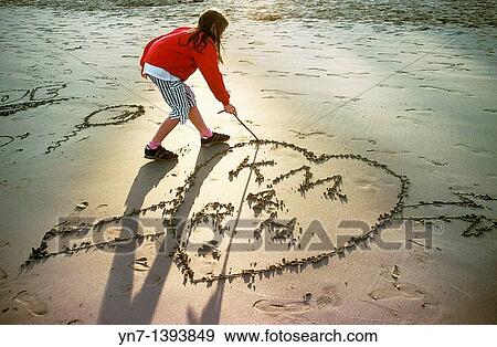 Young girl draws a love heart in the sand at the beach as the sun fades into an evening glow View Large Photo Image Stock Photo - Young girl draws a love heart in the sand at the beach as the sun fades into an evening glow. Fotosearch