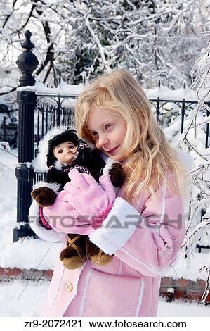 Stock Image - Young girl holding an eskimo doll outdoors near a snowy iron fence and frosted trees.. Fotosearch