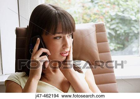 Picture - Young girl sitting curled up in a big arm chair by the window on the mobile looking serious. Fotosearch