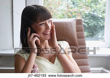 Stock Image - Young girl sitting curled up in a big arm chair by the window on the mobile smiling.. Fotosearch