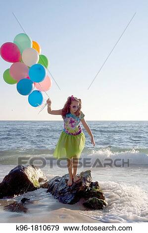 Stock Photo - young girl standing on rocks with balloons at the ocean. Fotosearch