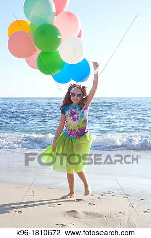 Stock Image - young girl walking towards camera on the beach holding balloons. Fotosearch