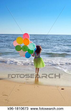 Stock Image - young girl walking towards the ocean with balloons with a view to see forever. Fotosearch