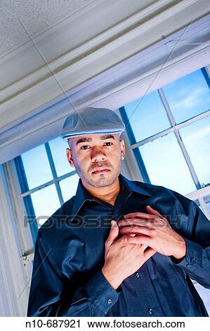 Stock Image - Young Hispanic man, wearing a cap on his head, posing in front of several windows.. Fotosearch