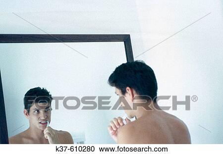 Stock Image - Young man brushing his teeth & looking in bathroom mirror. Fotosearch