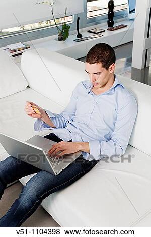 Young man buying in internet with credit card sitting on sofa View Large Photo Image Stock Photo - Young man buying in internet with credit card sitting on sofa. Fotosearch