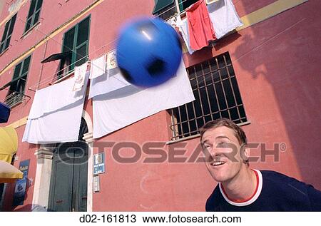 Stock Image - Young man heads blue soccer ball in front of clothes hanging out to dry in center of town, Vernazza, Cinqueterre, Italy. Fotosearch