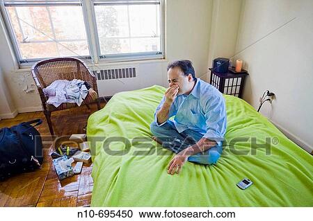 Young man, his cell phone beside him, sitting on his bed in his apartment. View Large Photo Image Stock Image - Young man, his cell phone beside him, sitting on his bed in his apartment.. Fotosearch