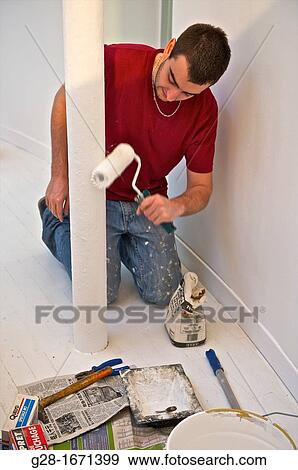 Stock Photo - Young man in red T-shirt and stained jeans using a paint roll to paint in white a pillar with tools around him. Fotosearch