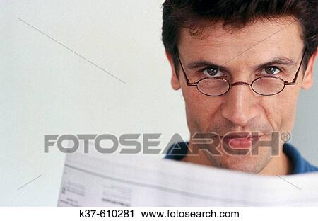 Stock Image - Young man looking over reading glasses holding newspaper. Fotosearch