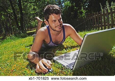 Young man lying on lawn and working with a laptop View Large Photo Image Stock Photography - Young man lying on lawn and working with a laptop. Fotosearch