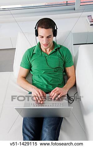 Stock Photography - Young man relaxed lying on the couch with laptop and listening to music. Fotosearch