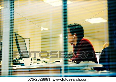 Young man seen through a window, working in an office in front of a computer View Large Photo Image Stock Image - Young man seen through a window, working in an office in front of a computer. Fotosearch