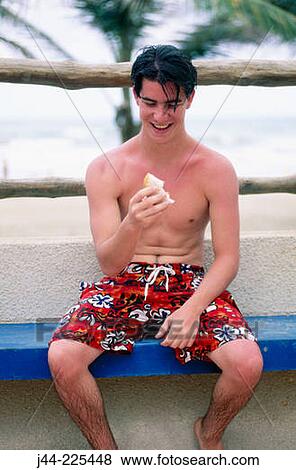 Stock Photo - Young man sitting on a bench eating a sandwich at the beach. Fotosearch