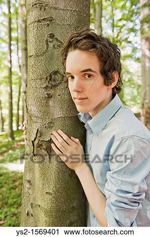 Young man standing by a tree in the woods View Large Photo Image Stock Image - Young man standing by a tree in the woods. Fotosearch