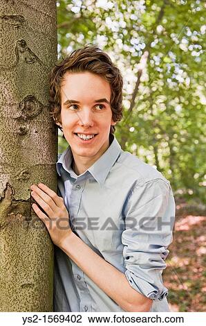 Young man standing by a tree in the woods View Large Photo Image Stock Image - Young man standing by a tree in the woods. Fotosearch