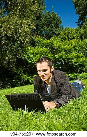 Young man working on his laptop computer in the garden, lying in the grass smiling View Large Photo Image Stock Photo - Young man working on his laptop computer in the garden, lying in the grass smiling. Fotosearch