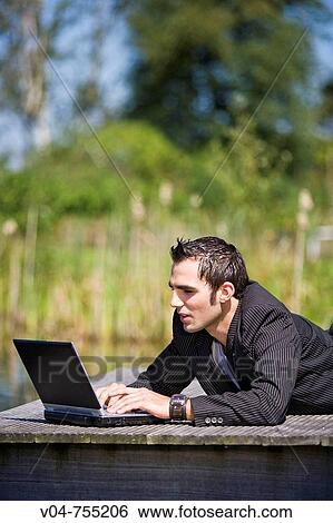 Young man working on his laptop computer lying on a footbridge View Large Photo Image Stock Photograph - Young man working on his laptop computer lying on a footbridge. Fotosearch
