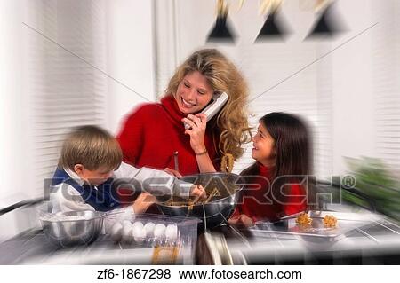 Young mother in her 30s on phone while she helps her two children make cookies in the home kitchen View Large Photo Image Stock Photo - Young mother in her 30s on phone while she helps her two children make cookies in the home kitchen. Fotosearch