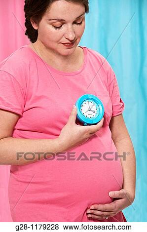 Young pregant woman in a pink top holding a blue alarm clock and her protruding belly View Large Photo Image Stock Photo - Young pregant woman in a pink top holding a blue alarm clock and her protruding belly. Fotosearch