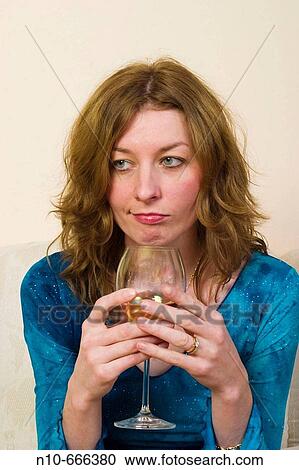 Stock Image - Young redhead woman sitting on her sofa, drinking a glass of wine.. Fotosearch