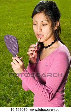 Stock Image - Young Spanish/Asian woman putting on makeup at a park.. Fotosearch