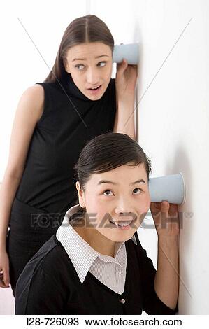 Stock Image - Young two businesswomen holding Disposable Cup against wall, listening. Fotosearch
