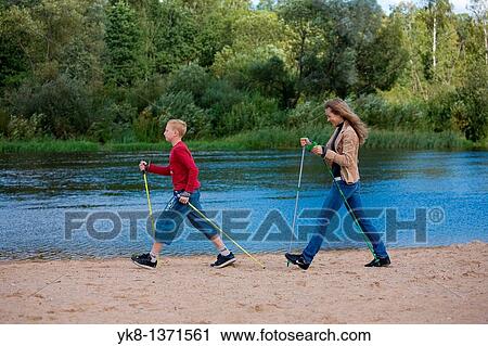 Stock Image - Young woman and teenage boy doing nordic walking by river water. Fotosearch
