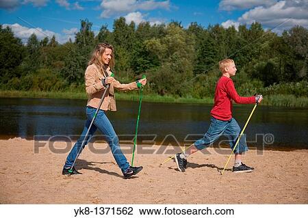 Stock Image - Young woman and teenage boy doing nordic walking by river water. Fotosearch