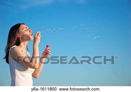 Stock Image - Young woman blowing bubbles against blue sky, close-up. Fotosearch