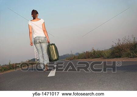 Stock Photo - Young woman carrying suitcases on road, rear view. Fotosearch