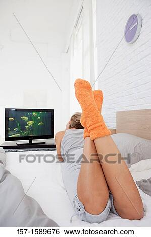 Stock Photograph - Young woman chilling out on her bed watching TV. Fotosearch