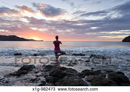 Stock Image - Young woman doing yoga at sunset in front of ocean surf at Playas del Coco, Costa Rica. Fotosearch