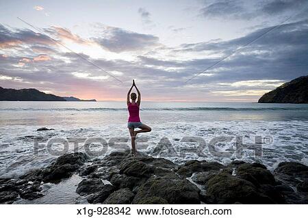 Stock Image - Young woman doing yoga on rocks at sunset in front of ocean surf at Playas del Coco, Costa Rica. Fotosearch