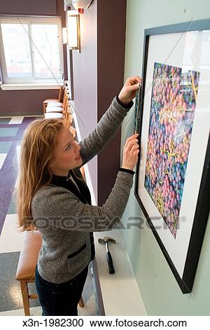 Stock Image - Young woman hanging photographs on wall.. Fotosearch