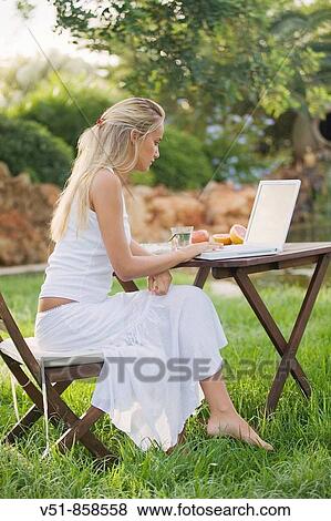 Stock Photo - Young woman having breakfast in the garden with laptop. Fotosearch