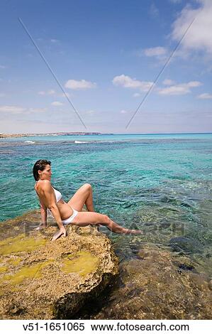 Stock Photography - Young woman in bikini sitting on the rocks and enjoying the sea in Formentera, Spain. Fotosearch