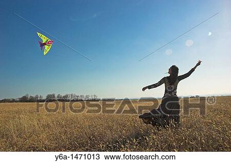 Stock Image - Young woman in field flying kite. Fotosearch